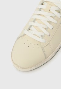 Αθλητικό παπούτσι από δέρμα κρεμ χρώματος με λευκά κορδόνια, διατρήσεις στην περιοχή των δαχτύλων και ελαφρώς φθαρμένη ελαστική σόλα σε λευκό φόντο.