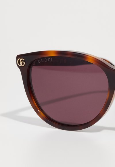 Slnečné okuliare Gucci s rámom z korytnačej škrupiny, fialovými šošovkami a zlatými logovými akcentmi; uhlový tvar s lesklým povrchom.
