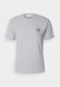 T-shirt gris clair à manches courtes avec un petit logo GANT comprenant un bouclier rouge, blanc et bleu et le texte "GANT 1949 NH CT" sur la poitrine.