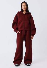 Sudadera burdeos con capucha y bolsillos delanteros, combinada con pantalones de pierna ancha. Textura suave y ajuste relajado. Zapatillas negras completan el conjunto.