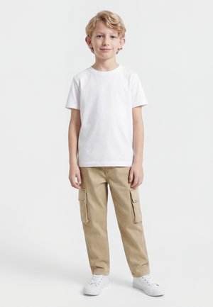 Blond gutt står, iført en enkel hvit t-skjorte, beige cargobukser og hvite joggesko mot en ensfarget hvit bakgrunn.