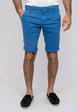 Homme portant un short bleu retroussé, des chaussures noires sans lacets et une chemise blanche, debout avec une main dans la poche sur un fond uni.