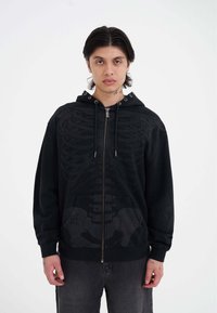 Sudadera negra con cremallera y diseño de esqueleto, que incluye capucha, bolsillos frontales y puños acanalados. El material parece suave y texturizado.