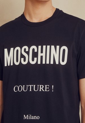 Person trägt ein schwarzes Moschino T-Shirt mit weißem Text "MOSCHINO COUTURE! Milano" auf der Vorderseite.