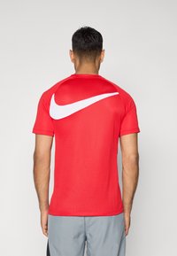 Piros Nike sportpóló rövid ujjakkal, texturált anyagból, és egy nagy fehér swoosh grafikával a hátsó részén.