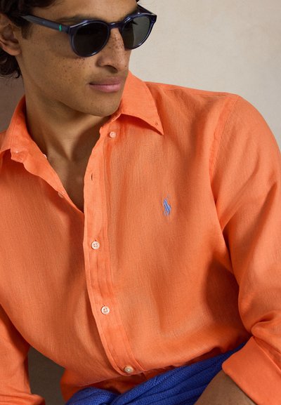 Άνδρας με σκουρόχρωμα γυαλιά ηλίου και πορτοκαλί πουκάμισο με κουμπιά και μπλε κεντητό λογότυπο στο στήθος, με μπλε ύφασμα τοποθετημένο πάνω στη λεκάνη.