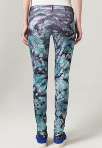 Jean skinny avec un motif tie-dye dans des teintes de bleu-vert et noir, comportant cinq poches et des détails de couture. Texture en denim douce.