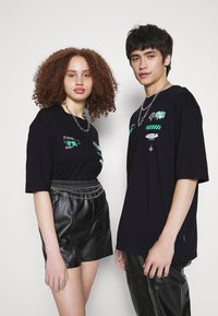 Černé oversized trička s grafickými akcenty; jeden model má na sobě stříbrný řetězový pás s koženými šortkami, druhý v kožených kalhotách.