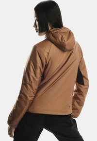 Femeie văzută din spate, purtând o jachetă cu glugă de culoare bej deschis și pantaloni negri, uitându-se pe lateral pe un fundal alb simplu.