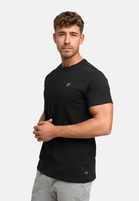 Black cotton t-shirt with short sleeves, round neckline, and a small white logo at the chest. Model stands with arms crossed, wearing light gray pants.