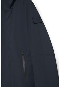 Mörkblå jacka med en slät textur, som har ett märkespatch på axeln och en dragkedjeficka på sidan.