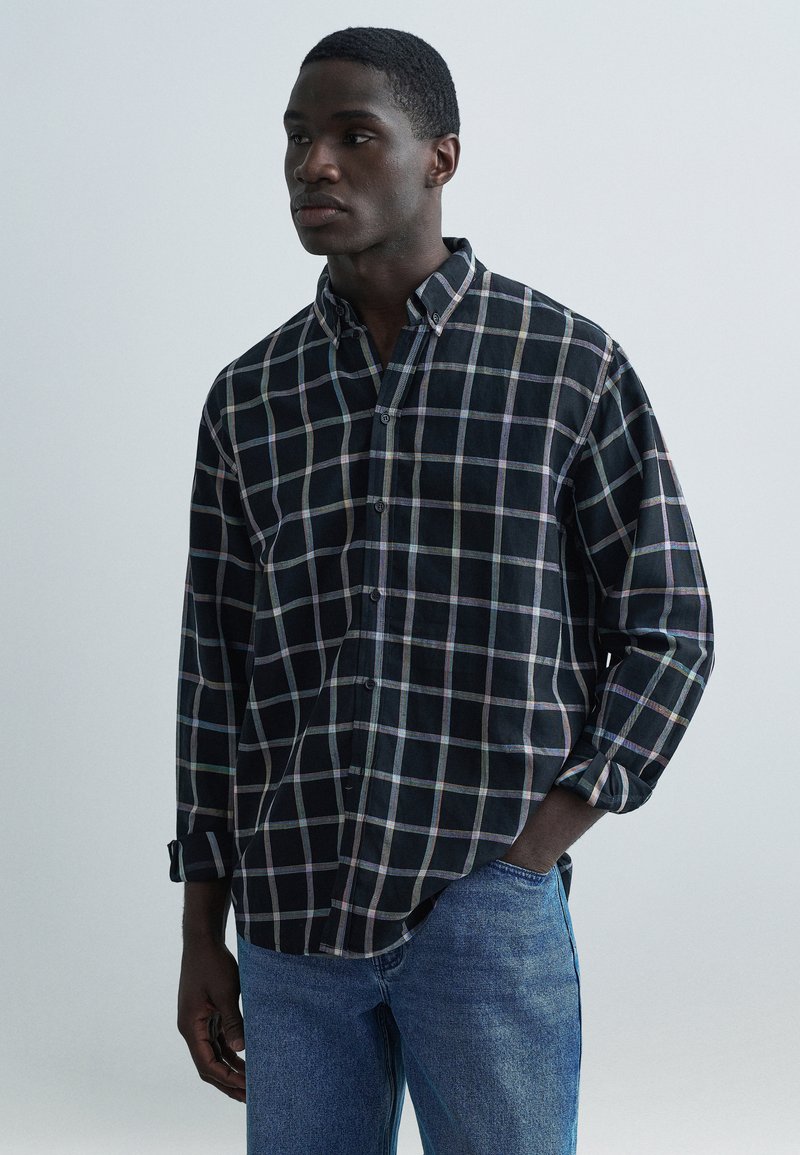 Μαύρο και πολύχρωμο καρό πουκάμισο με γυρισμένα μανίκια, γιακά με κουμπιά και απαλό ύφασμα· συνδυάζεται με μπλε τζιν τζιν.