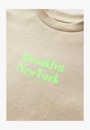 Béžová mikina s jemným okrajom na krku a jasne zeleným textom "Brooklyn New York" s nápisom "Premium Goods Supply" pod ním.