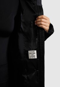 Zwarte geïsoleerde jas met een gladde buitenkant, voorzien van een ritssluiting aan de zak en een label met de tekst "Een warme omhelzing gemaakt van stof."