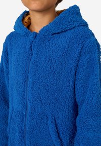 Modra flisasta zip-up jopica z teksturirano površino, velika kapuca in žepi. Notrajnost obleke v kontrastni barvi. Mehka, udobna tkanina.