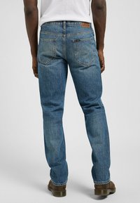 Jeans de denim azul, de pierna recta, de talle medio, con un parche de cuero en la cintura, un canesú en la parte trasera y dos bolsillos traseros con detalles de costura.