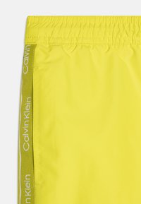 Shorts de bain jaunes vifs en matériau léger, dotés d'une taille élastique, d'une poche latérale et du logo Calvin Klein sur un accent de taille verte.