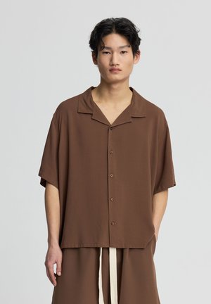 Giovane uomo con capelli neri corti che indossa una camicia marrone larga a maniche corte con bottoni e pantaloni marroni abbinati con coulisse bianca.