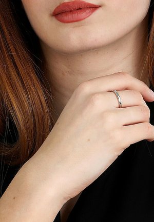 Nærbillede af en person med lys hud, rødt hår, der bærer rød læbestift, og holder hånden tættere på ansigtet og viser en simpel sølvring på fingeren.