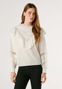 Vit blus med volanger i halsringningen och ärmarna, med broderi och spetsdetaljer. Mjuk tyg, avslappnad passform och svarta jeans.