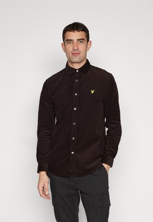 Camisa de pana marrón oscuro con cuello abotonado y un pequeño logotipo de águila amarilla en el pecho, con un diseño ajustado y mangas largas.