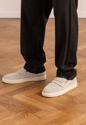Light gray slip-on sneakers with white soles worn with black pants on a wooden parquet floor.
