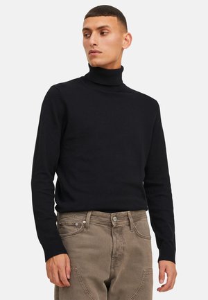 Mann trägt einen schwarzen Rollkragenpullover und hellbraune Jeans, steht und schaut seitlich vor einem weißen Hintergrund.