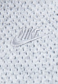 Nike-logotypen i broderad grå på en ljusgrå, strukturerad tyg med små perforeringar. Nätt närbild som framhäver material och detaljer.
