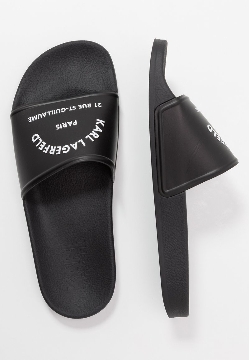 Des sandales à glissière noires avec une sangle supérieure texturée arborant un logo blanc. La semelle est plate et lisse, conçue pour le confort et la facilité de port.
