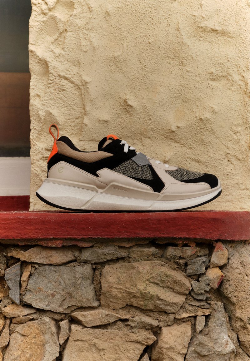 Sneaker mit einem oberen Teil aus beigem und schwarzem Mesh, orangefarbenen Akzenten, weißen Schnürsenkeln und einer klobigen weißen Sohle. Verfügt über strukturierte Details an den Seiten.