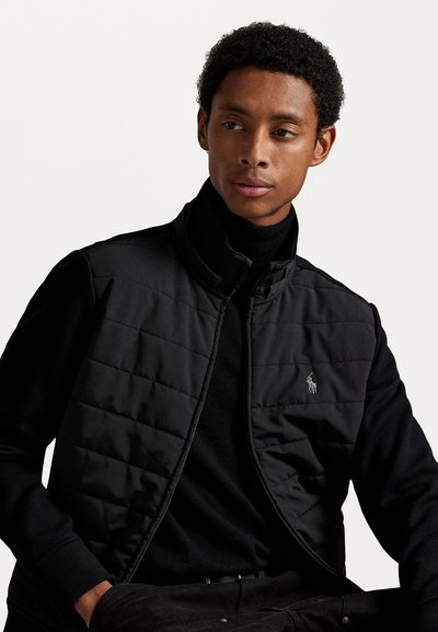 Μαύρο πλεκτό γιλέκο με ψηλό γιακά, που διαθέτει μια διακριτική λογότυπο. Φορεμένο πάνω από μια μαύρη μπλούζα με φίδια, αναδεικνύει μια λεία υφή υφάσματος.