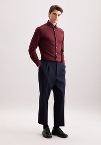 Camicia bordeaux con bottoni e maniche lunghe, abbinata a pantaloni a pieghe blu scuro e scarpe nere stringate; tessuto morbido, design aderente.