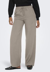 Donna che indossa pantaloni ampi beige con vita regolabile, mule flat neri con borchie e un top nero corto a maniche lunghe.