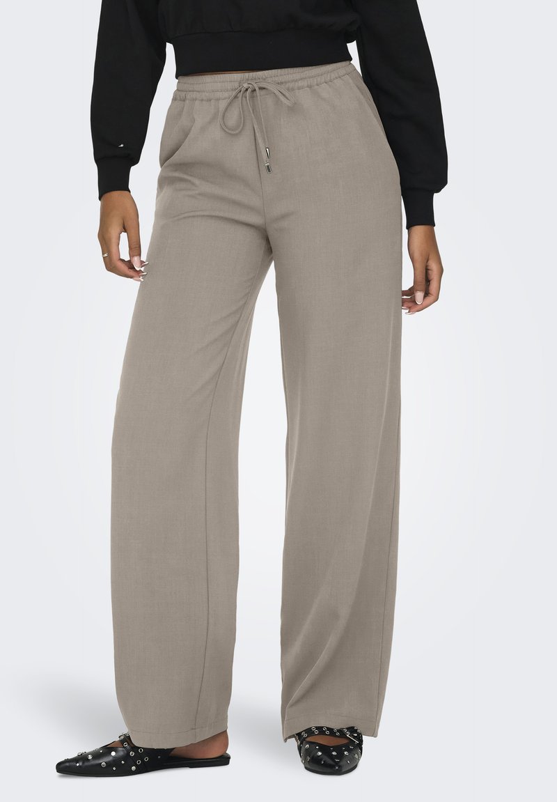 Donna che indossa pantaloni ampi beige con vita regolabile, mule flat neri con borchie e un top nero corto a maniche lunghe.