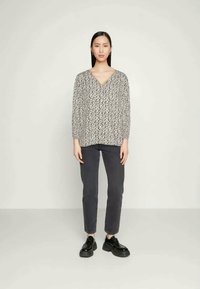 Donna con capelli lunghi che indossa una blusa a V nera e bianca con stampa floreale, jeans grigio scuro e scarpe con plateau nere, in piedi su uno sfondo semplice.