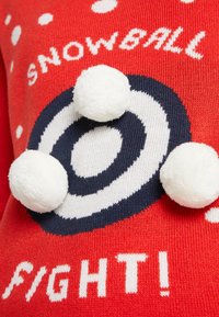 Pull en tricot rouge avec pois blancs et un motif circulaire bleu marine. Trois pompons blancs sont attachés, avec le texte "BATAILLE DE BOULES DE NEIGE !".