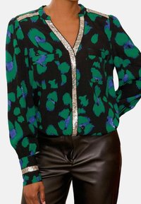Blusa negra con manchas abstractas en verde y azul, escote en V, dos bolsillos frontales y acentos de lentejuelas plateadas en los hombros y puños.
