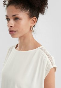 Blusa bianca in tessuto morbido, con scollo rotondo e maniche corte. Dettaglio in pizzo decorativo lungo la spalla che aggiunge texture.
