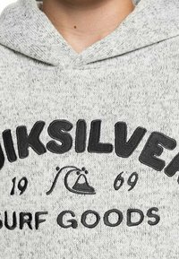 Primo piano di una felpa grigia a maglia con il testo ricamato in nero "QUIKSILVER 1969 SURF GOODS" e un logo con onda e montagna.