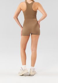 Brązowy, prążkowany zestaw sportowy składający się z crop topu i szortów. Model nosi jasnego kolorystycznie sneakersy z białymi skarpetkami. Tło jest jednolicie szare.