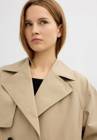 Trench coat beige con un amplio cuello, con una textura suave y detalle de botones, usado sobre una blusa negra de canalé.