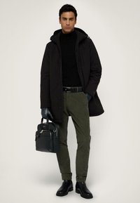 Abrigo negro con capucha sobre un suéter negro de cuello alto, pantalones verde oliva, guantes negros y botas negras hasta el tobillo. Un bolso verde oscuro con herrajes plateados.