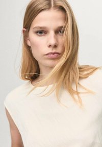 Una blusa sin mangas de color beige claro con una textura suave, sobre un fondo neutro. La modelo tiene el cabello lacio y rubio.