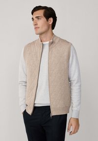 Pamučni tan prsluk s visokim kragnom, pletivnim otvorima za ruke i zatvaračem, složen preko svijetlosive majice s kapuljačom i tamnih hlača.