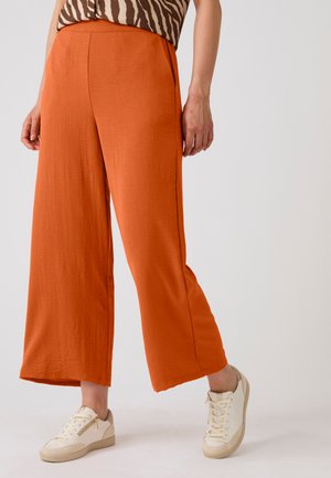 Donna che indossa pantaloni arancioni ampi e corti e sneakers crema, in piedi davanti a uno sfondo semplice.