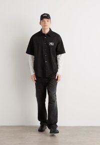 Zwarte shirt met korte mouwen met een knoopsluiting en een logo-patch, gedragen over een wit shirt met lange mouwen en gecombineerd met donkere broek.