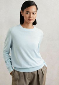 Helles blaues Sweatshirt aus weichem Material, mit langen Ärmeln, rundem Hals und enganliegendem Bund, kombiniert mit hellbraunen, plissierten Hosen.
