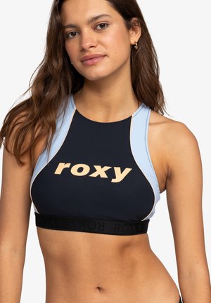 Sportowy biustonosz Roxy w kolorowym wzorze z granatowym i jasnoniebieskim, dużym nadrukiem logo oraz czarną elastyczną taśmą z logo. Gładka tekstura.
