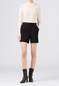 Schwarze maßgeschneiderte Shorts mit hohem Bund und Taschen, kombiniert mit einem cremefarbenen Langarmoberteil und schwarzen Stiefeletten mit Blockabsätzen.