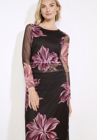 Žena oblečená v černých přiléhavých šatech s průhlednými dlouhými rukávy a velkými růžovými květinovými vzory, stojící proti jednoduchému bílému pozadí.